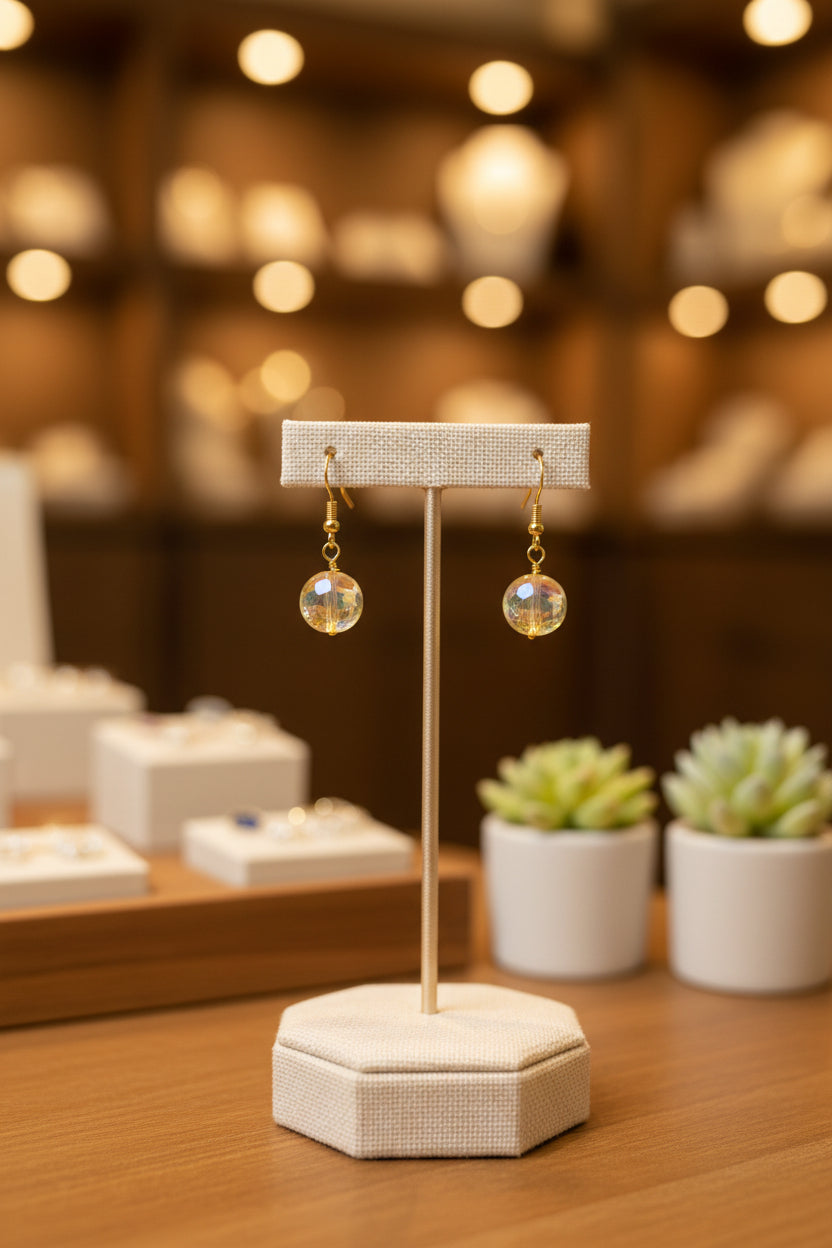 A pair of gold-tone dangle earrings with round, iridescent opal-like beads, displayed on a clear acrylic stand against a neutral gray background.