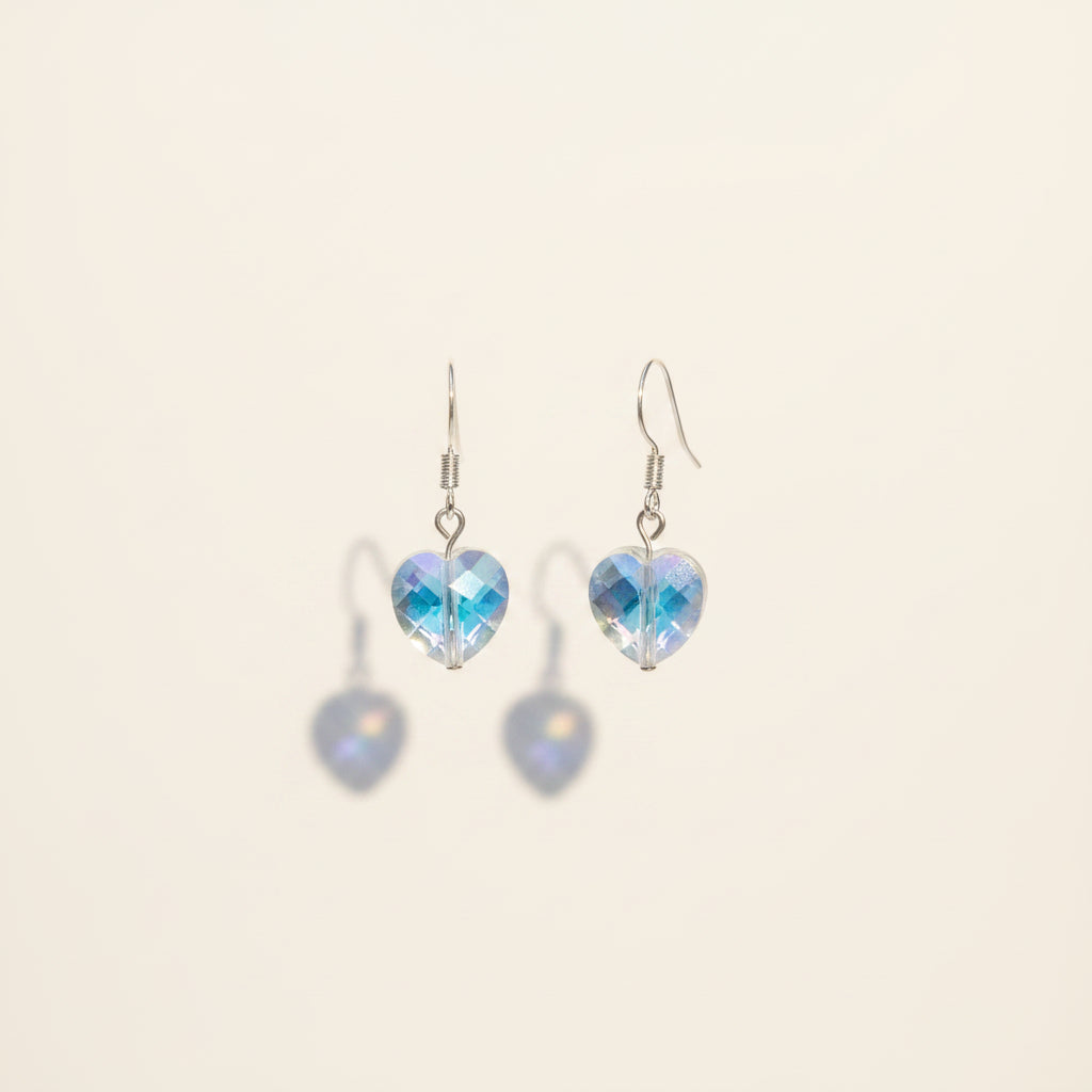 A pair of dangling heart-shaped earrings with iridescent blue and purple faceted stones on silver hooks, displayed against a neutral gray background.