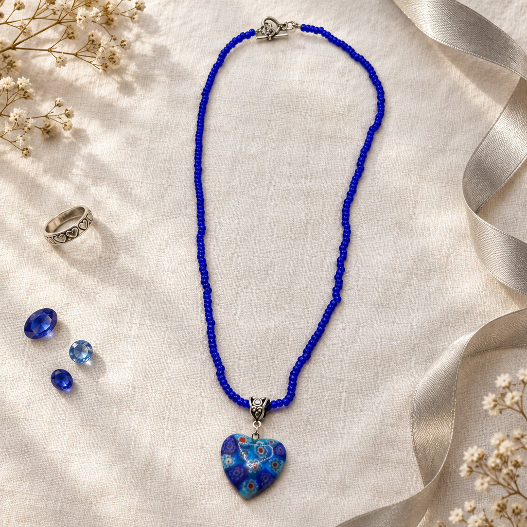 A beaded necklace with a heart-shaped glass pendant, featuring a vibrant blue cord and a blue and white patterned heart pendant on a light gray background.