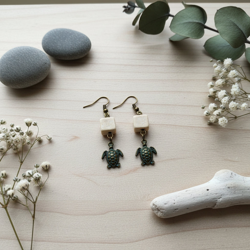 A pair of handmade dangle earrings featuring small cream-colored square beads and dangling sea turtle charms with blue and gold enamel detailing, displayed on a clear stand against a light gray background.