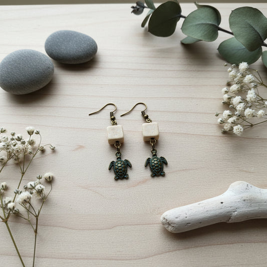 A pair of handmade dangle earrings featuring small cream-colored square beads and dangling sea turtle charms with blue and gold enamel detailing, displayed on a clear stand against a light gray background.