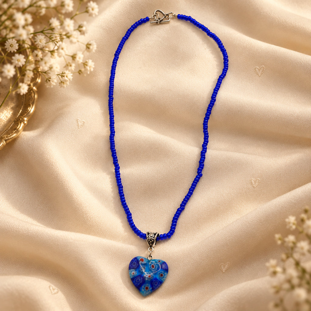 A beaded necklace with a heart-shaped glass pendant, featuring a vibrant blue beaded chain and a blue glass heart pendant with red and white patterns, set against a plain light gray background.