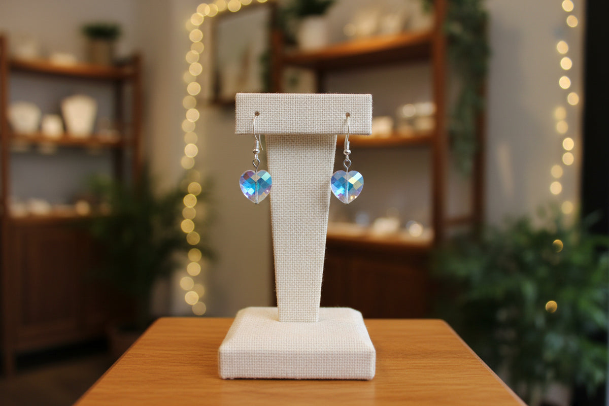A pair of silver-tone dangle earrings with faceted heart-shaped crystals that have an iridescent blue and purple sheen, displayed on a clear acrylic stand against a neutral gray background.