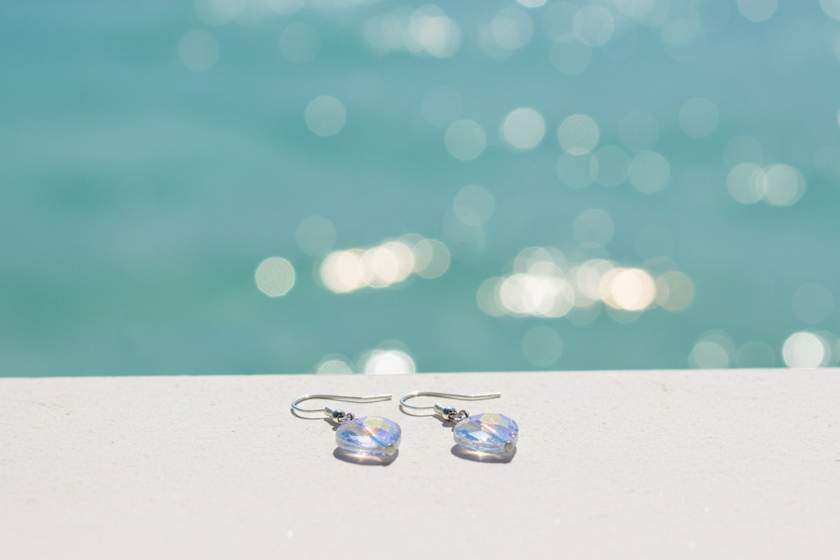A pair of heart-shaped dangling earrings with iridescent blue and purple faceted crystal pendants on silver hooks, displayed on a clear stand against a plain gray background.