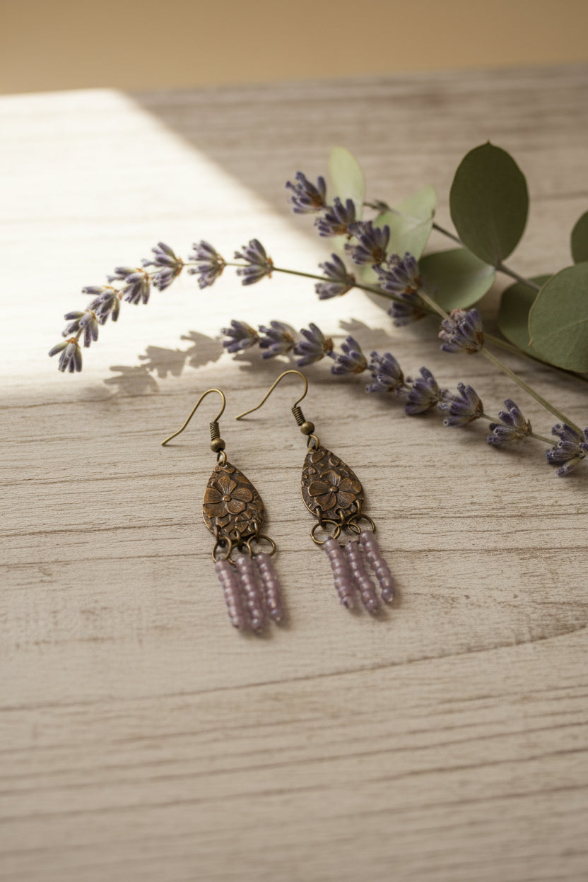 A pair of dangling earrings with textured bronze teardrop-shaped metal tops, small silver rings, and pink beaded fringe, displayed against a plain white background.