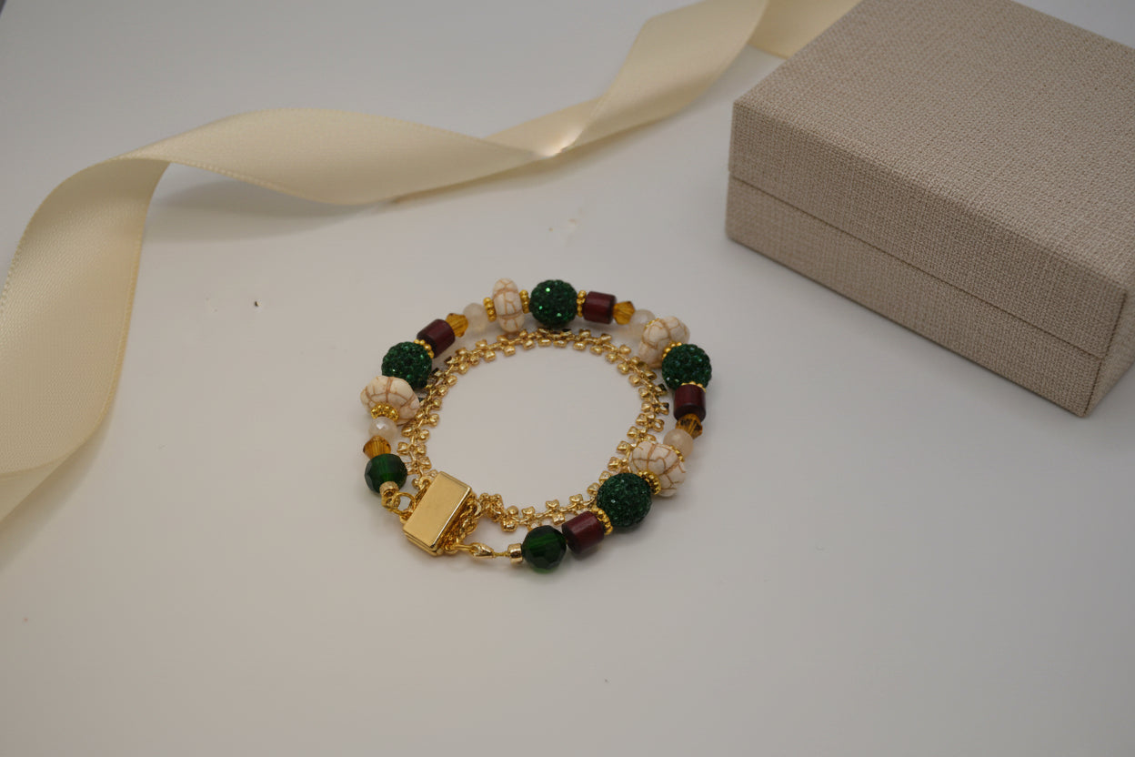 A gold-tone beaded bracelet with dark green textured beads, cream-colored beads, and small maroon accents, displayed on a plain light gray background.
