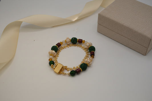 A gold-tone beaded bracelet with dark green textured beads, cream-colored beads, and small maroon accents, displayed on a plain light gray background.