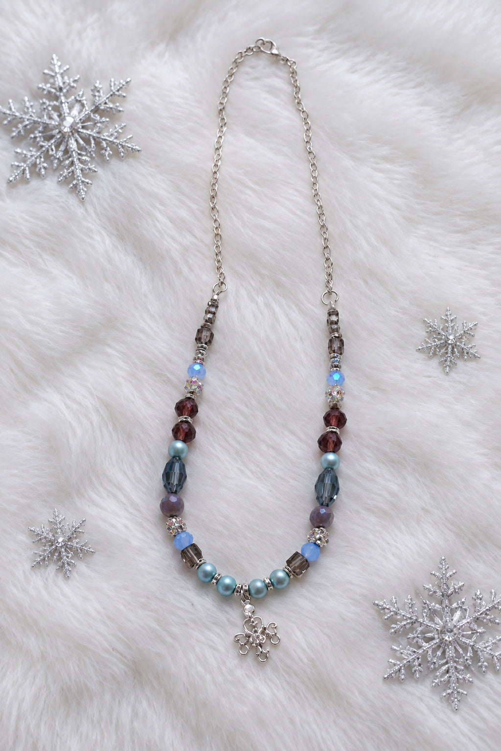 A beaded necklace featuring light blue, purple, and reddish-brown beads with a silver-toned snowflake charm, displayed on a plain light gray background.