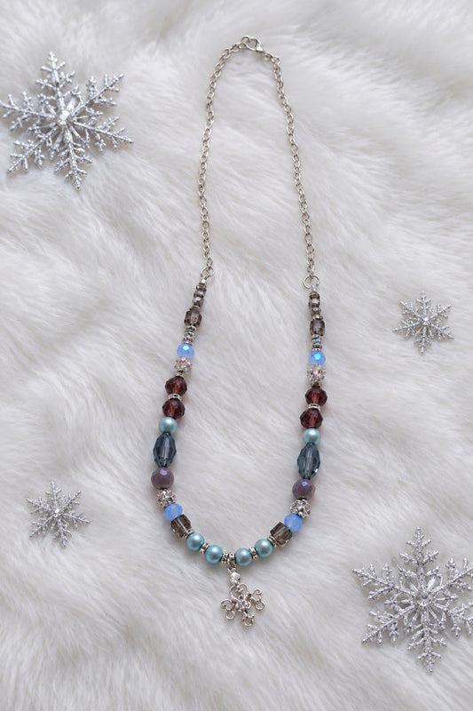 A beaded necklace featuring light blue, purple, and reddish-brown beads with a silver-toned snowflake charm, displayed on a plain light gray background.