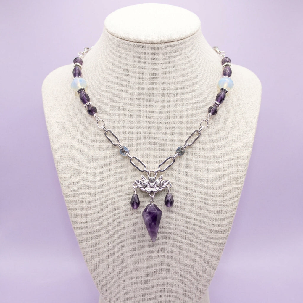 A silver-toned beaded necklace with a bat-shaped pendant and purple crystal drops, displayed on a black velvet jewelry bust.