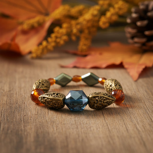 A beaded stretch bracelet featuring ornate gold filigree oval beads, translucent amber-orange round beads, two square sage green beads, and a faceted dark blue-green center bead, displayed on a neutral light gray background.