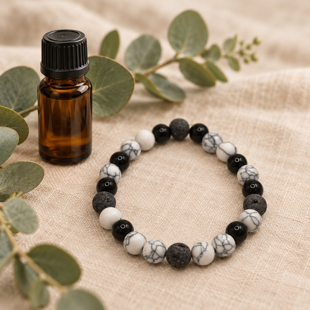 A beaded stretch bracelet made of black, white, and marbled gray beads, styled on a beige linen fabric next to an amber glass bottle and green eucalyptus leaves.