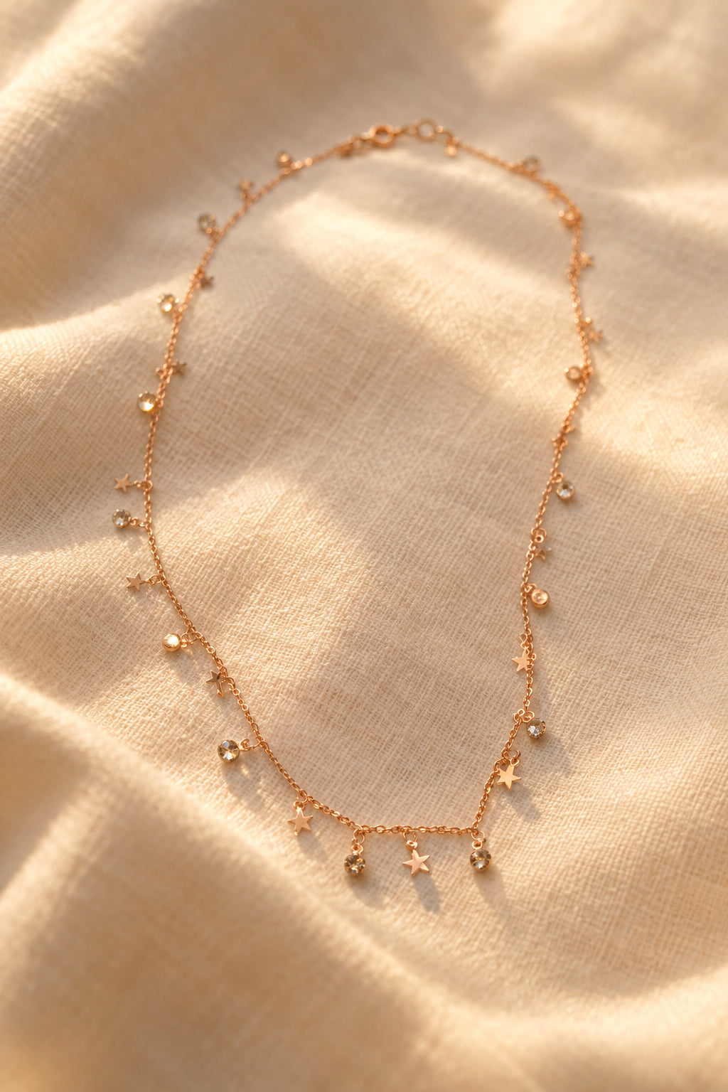 A delicate rose gold necklace with small dangling charms, including stars and crescent moons, laid out on a plain gray background.