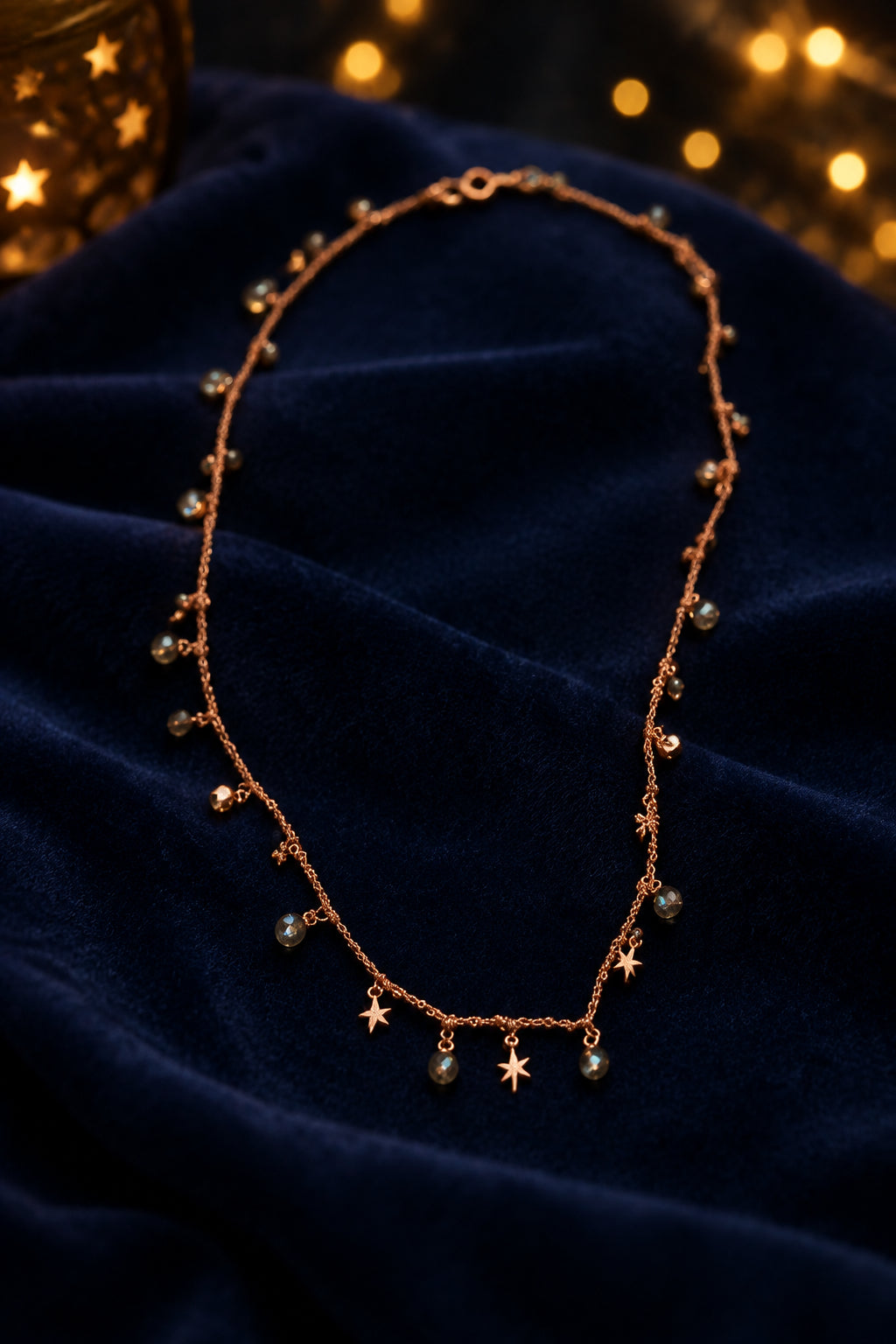 A delicate rose gold necklace with small dangling charms, including stars and crescent moons, laid flat on a neutral gray background.