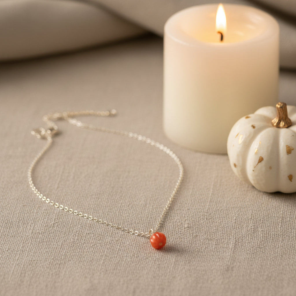 A delicate gold chain necklace with a small round orange pendant, displayed on a black velvet jewelry bust against a white background.