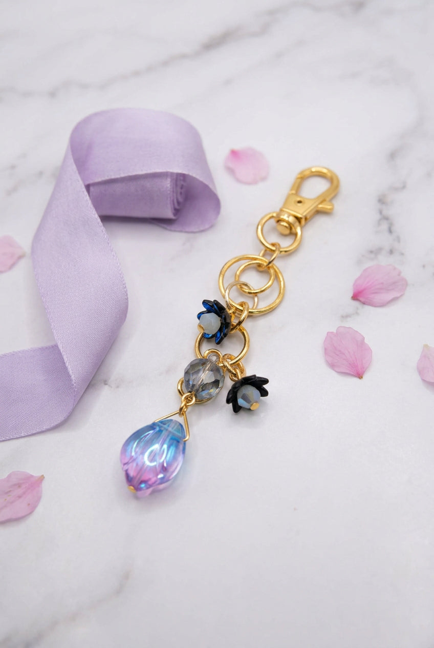 A gold-toned lobster clasp earring with a dangling charm featuring black bat wings, a gray stone, and a faceted iridescent purple-blue bead, displayed on a clear acrylic stand against a white background.