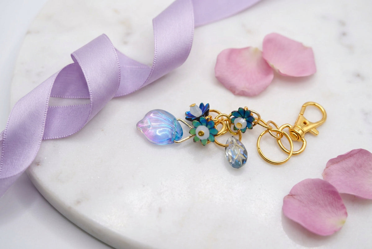 A gold-tone metal keychain charm featuring a cluster of colorful glass beads and flower-shaped charms, including a prominent iridescent purple and blue bead, set against a plain light gray background.