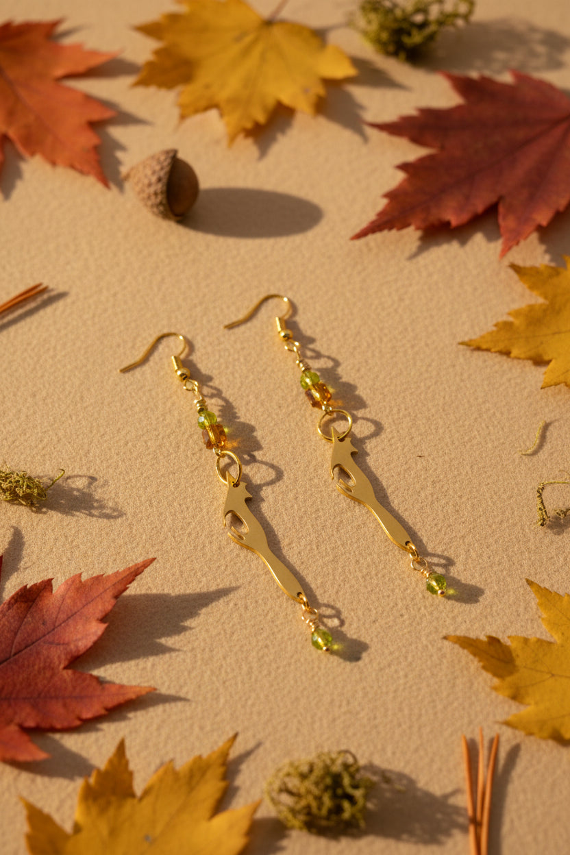 A pair of gold-toned dangle earrings with green faceted beads and a small dangling green bead, displayed against a light gray arched backdrop.