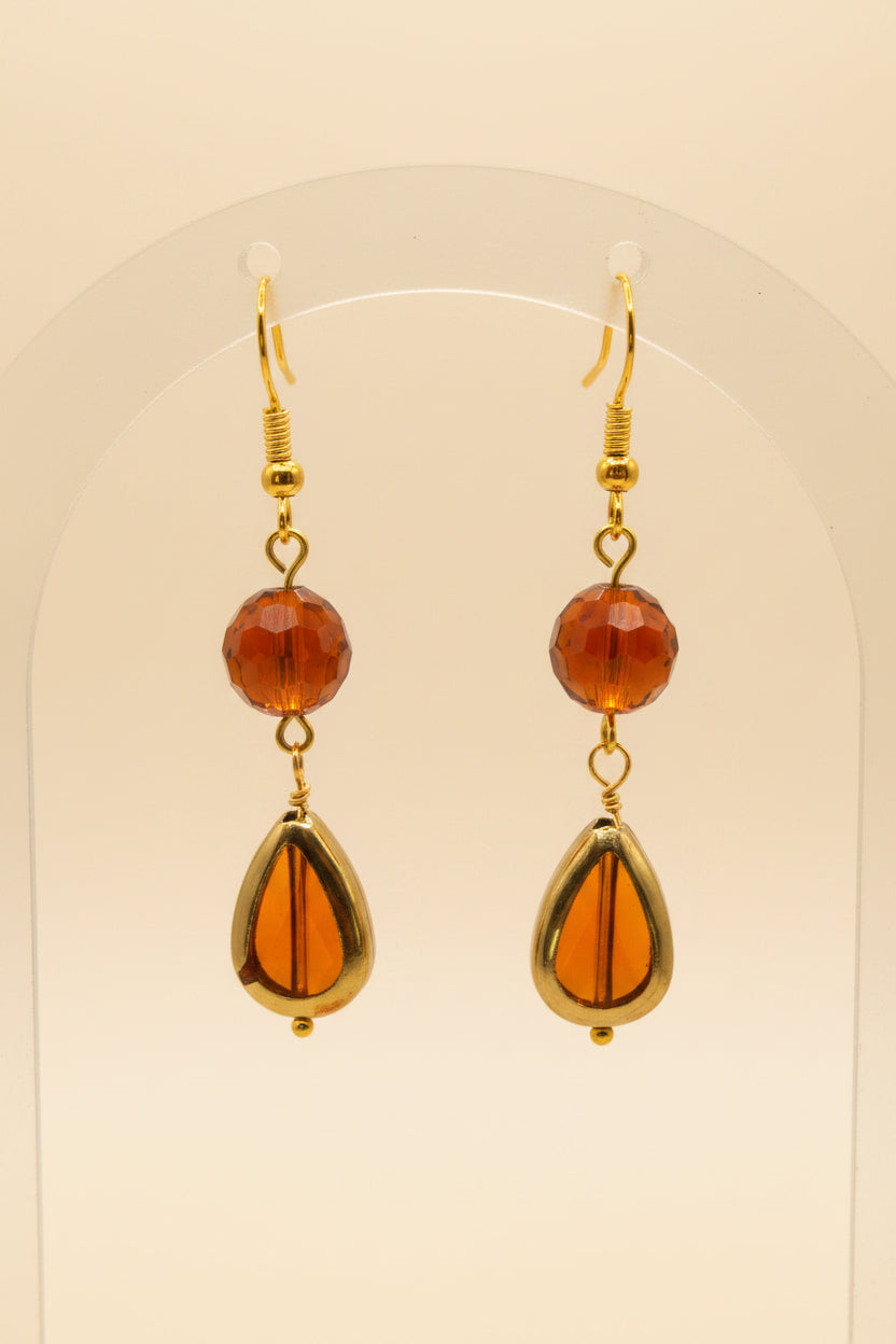 A pair of gold-tone dangle earrings with round amber-brown beads and teardrop-shaped amber drops, displayed on a clear stand against a light gray background.