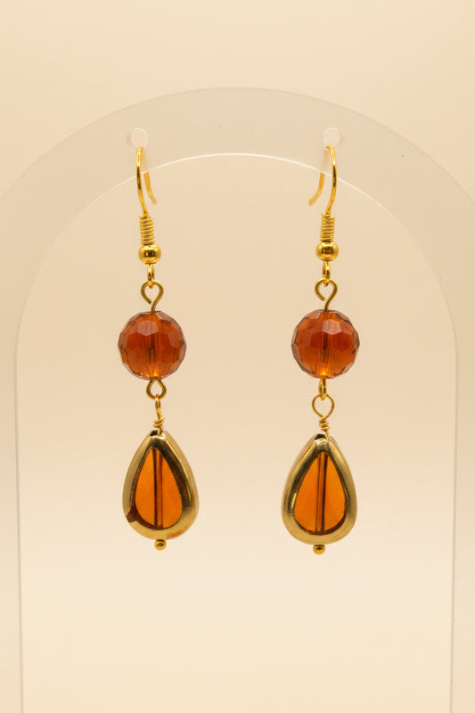 A pair of gold-tone dangle earrings with round amber-brown beads and teardrop-shaped amber drops, displayed on a clear stand against a light gray background.