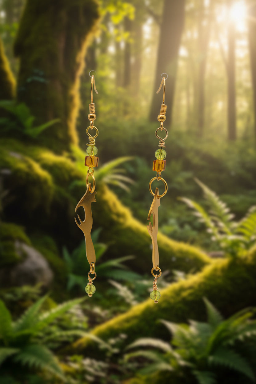 A pair of gold-toned drop earrings with bottle opener charms, small amber beads, and light green faceted beads, hanging against a white arched display backdrop.