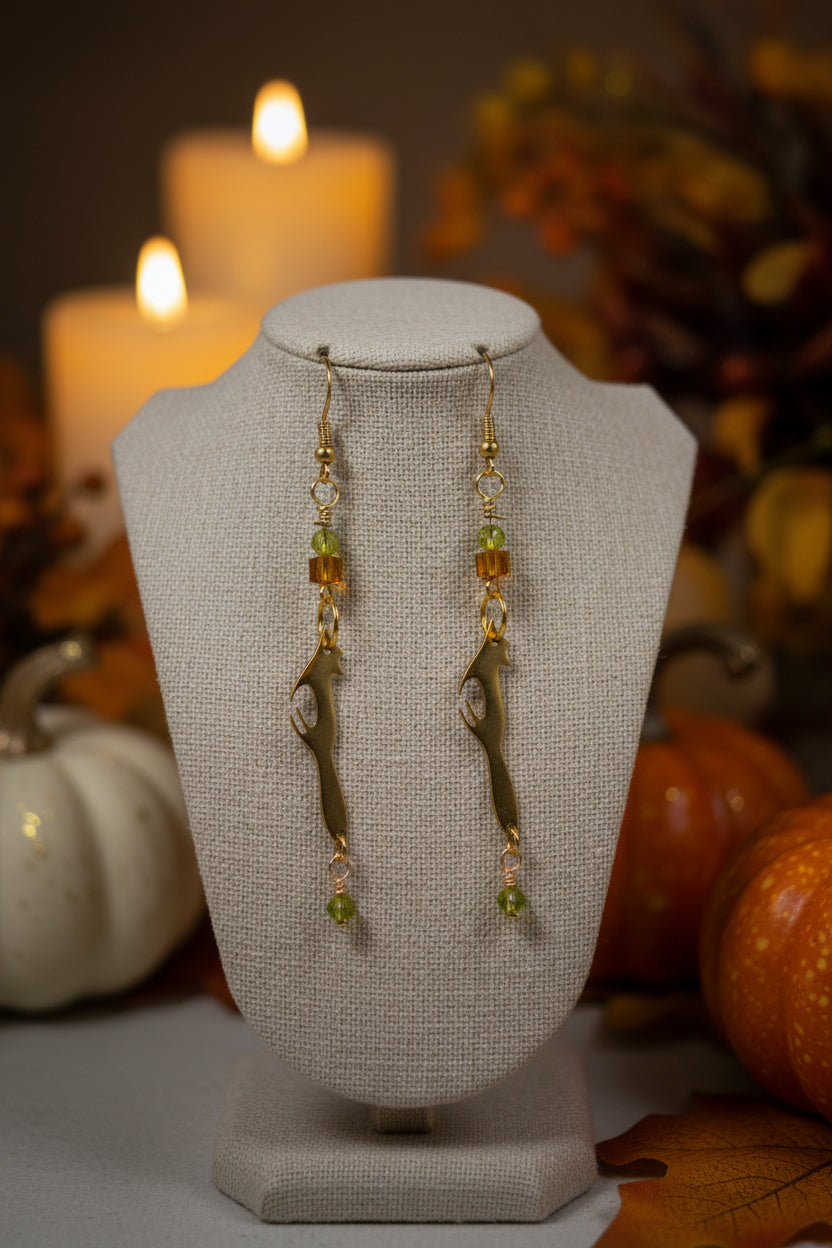 A pair of gold-toned dangle earrings with fox-shaped charms, green faceted beads, and small amber cube beads, displayed against a white arched backdrop.