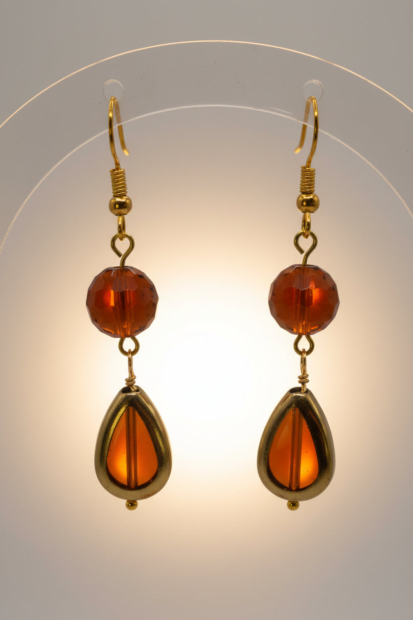 A pair of gold-tone dangle earrings with round amber-brown beads and teardrop-shaped amber drops, displayed on a clear stand against a light gray background.
