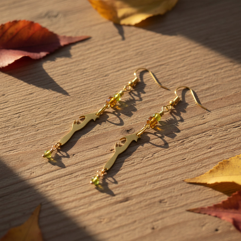 A pair of gold-toned dangle earrings displayed on a clear plastic stand against a light gray background. The earrings feature a stylized cat silhouette charm, small green faceted beads, and amber-colored beads.