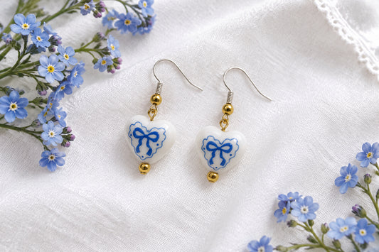 A pair of dangling heart-shaped earrings with white enamel, blue bow designs, and small gold-toned metal accents, displayed against a light gray background.