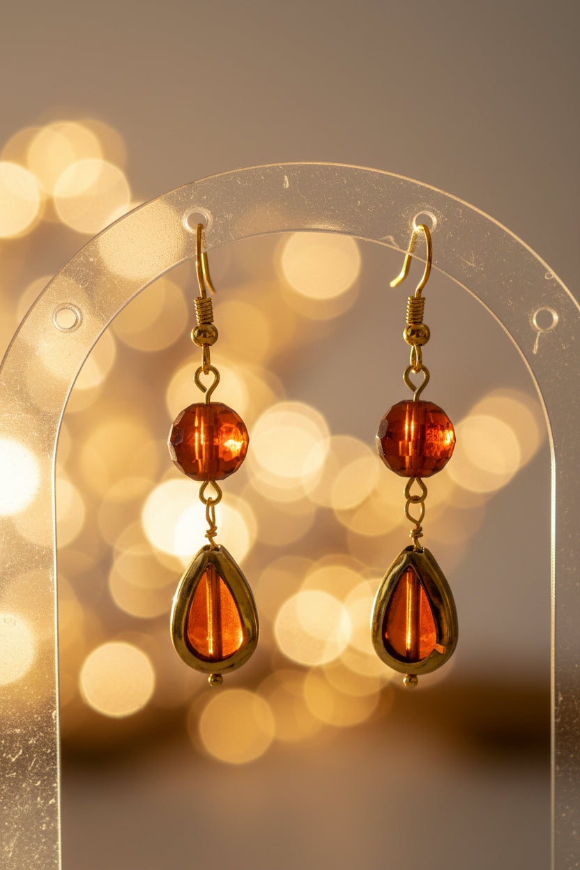 A pair of gold-tone dangle earrings with round amber-brown beads and teardrop-shaped amber drops, displayed against a light gray background.