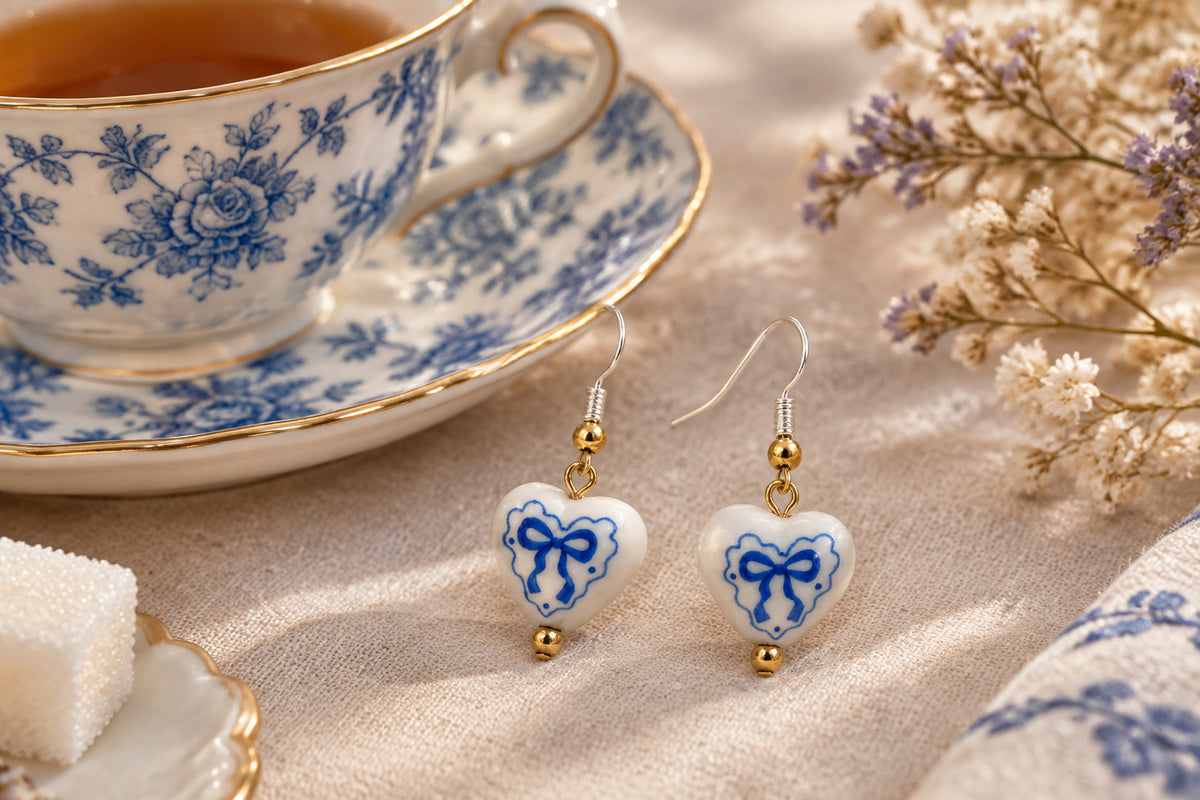 A pair of dangling earrings with small white heart-shaped beads decorated with blue bow motifs, suspended from silver hooks with gold-toned beads and bells, set against a neutral gray background.