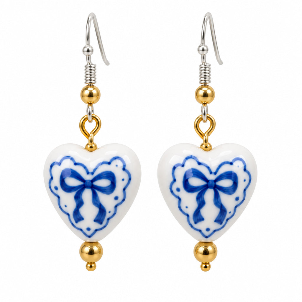 A pair of dangling heart-shaped earrings with white enamel or porcelain fronts decorated with blue bow motifs, hanging from gold-toned hooks against a plain light gray background.