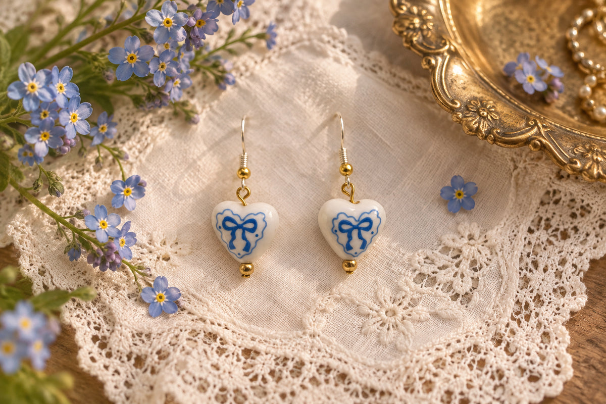 A pair of dangling earrings with small white heart charms printed with a blue bow design, hanging from silver hooks with gold-toned beads on a clear display stand against a light gray background.