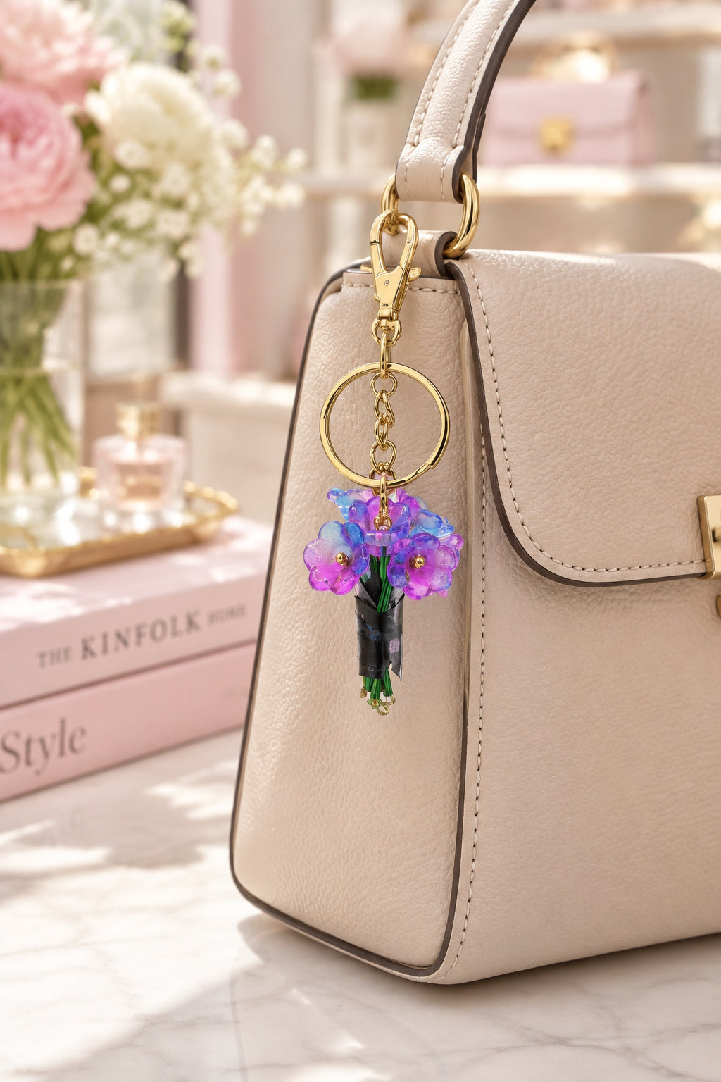 A small gold-tone keychain with a floral charm featuring purple and blue flowers, displayed on a clear acrylic stand with a wooden base against a light gray background.