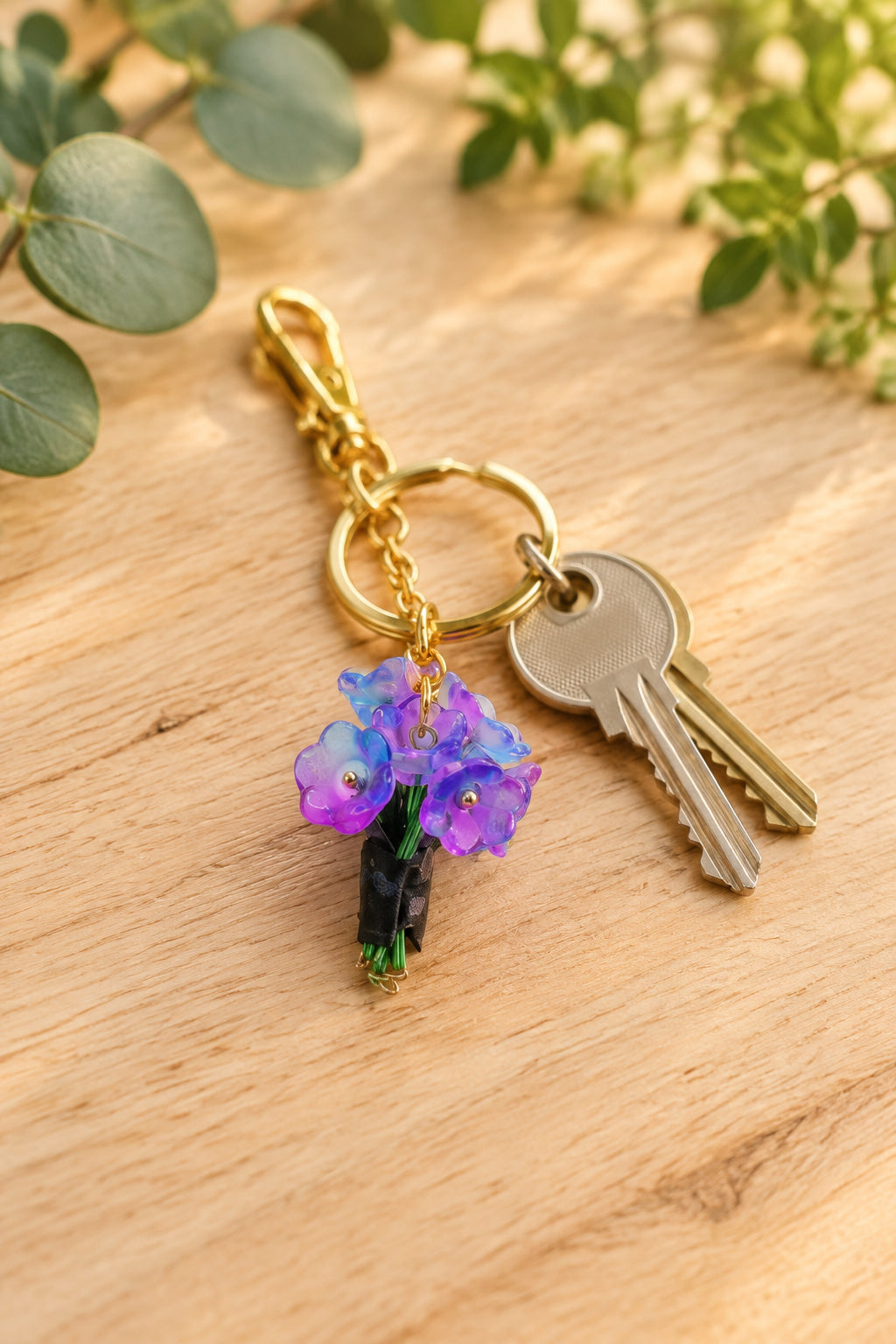 A gold-tone keychain with purple and blue flower charms hanging from a clear acrylic display stand on a light gray background.