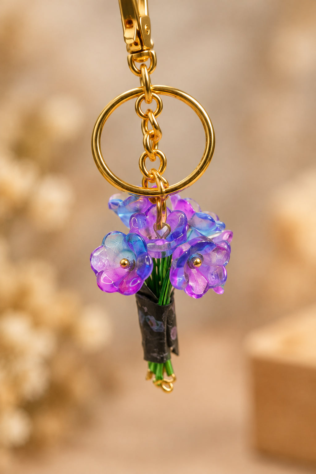 A gold-tone keychain with a small purple and blue flower charm hanging from a gold ring, displayed on a clear acrylic stand with a wooden base against a light gray background.