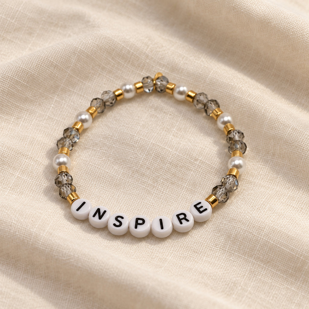 A beaded stretch bracelet featuring white letter beads spelling 'INSPIRE' with black text, surrounded by clear faceted beads and small gold-toned spacer beads, set against a plain light gray background.