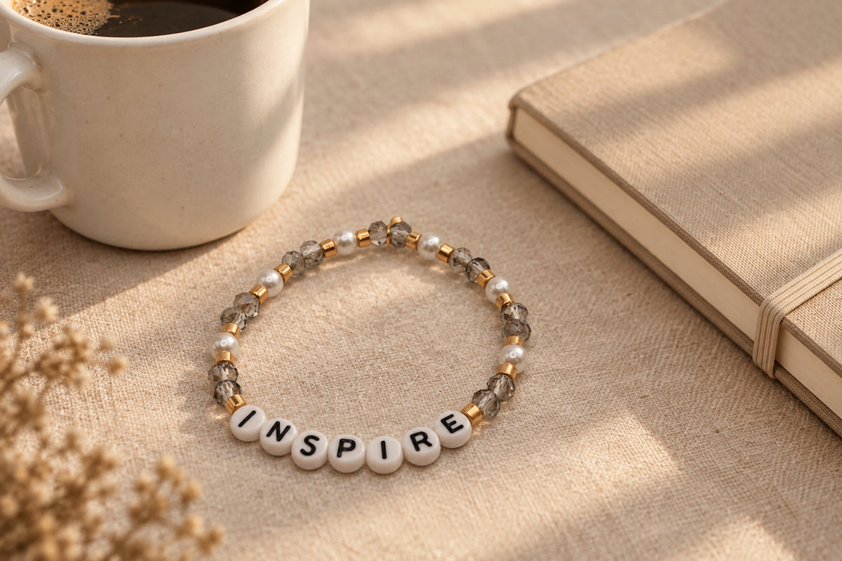 A beaded stretch bracelet on a neutral gray background, featuring white letter beads spelling 'INSPIRE' with black lettering, surrounded by clear faceted beads and small gold spacer beads.