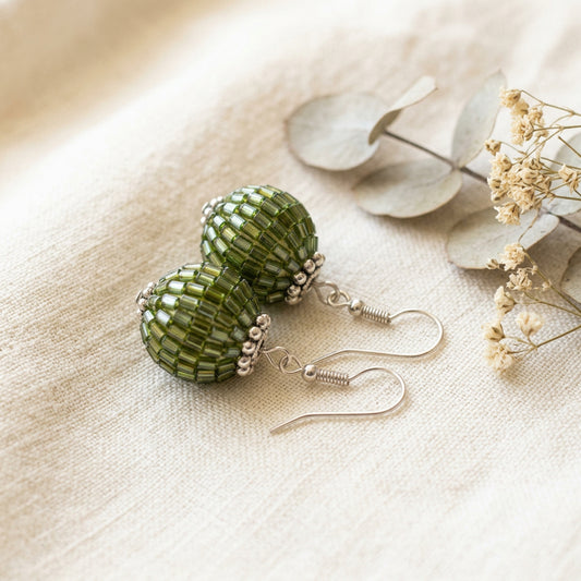A pair of handmade dangle earrings with textured, ribbed green glass beads and silver hooks, displayed on a clear acrylic stand against a neutral gray background.