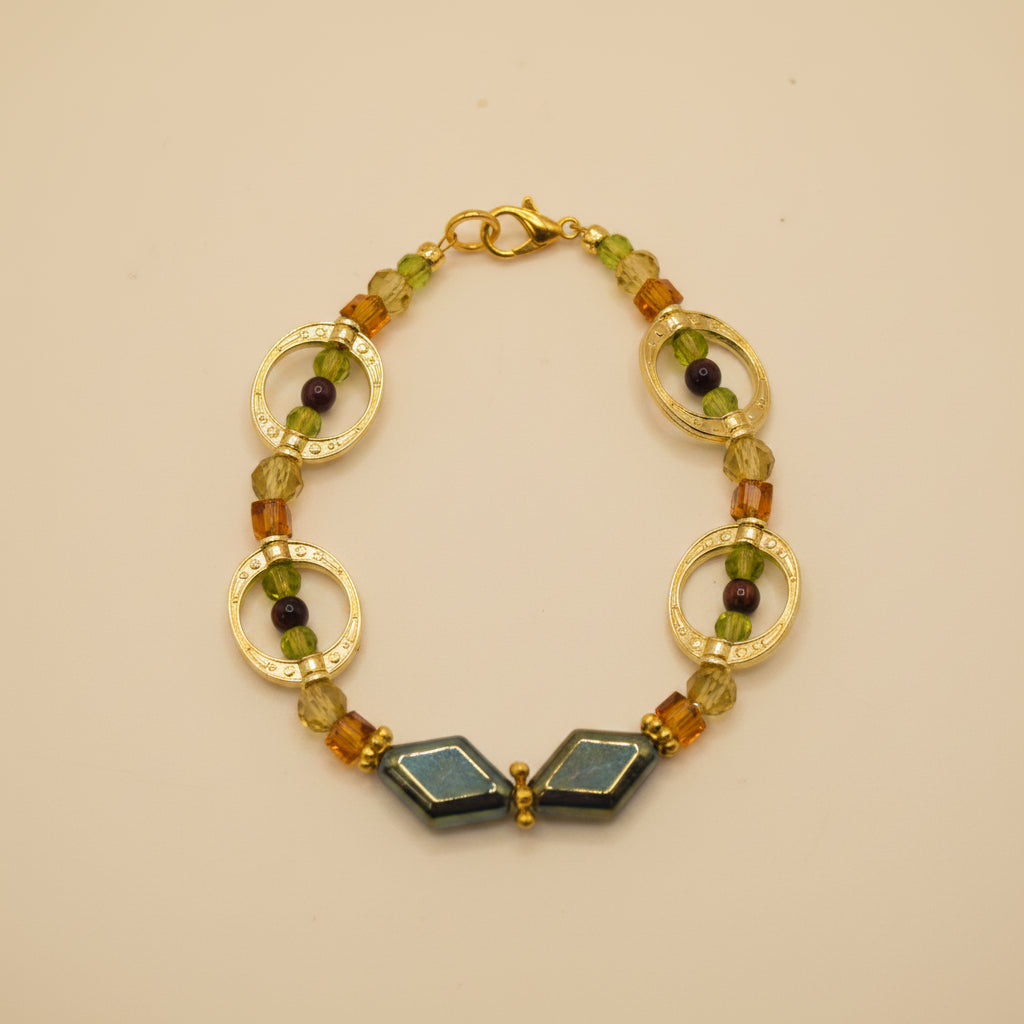 A gold-tone beaded bracelet featuring two large textured circular links, small amber and olive green beads, and two dark green faceted stone beads at the bottom, displayed on a neutral gray background.