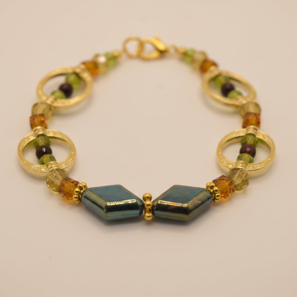 A gold-tone beaded bracelet featuring circular gold spacer rings, faceted amber and green beads, and two green diamond-shaped beads at the front, displayed on a neutral light gray background.