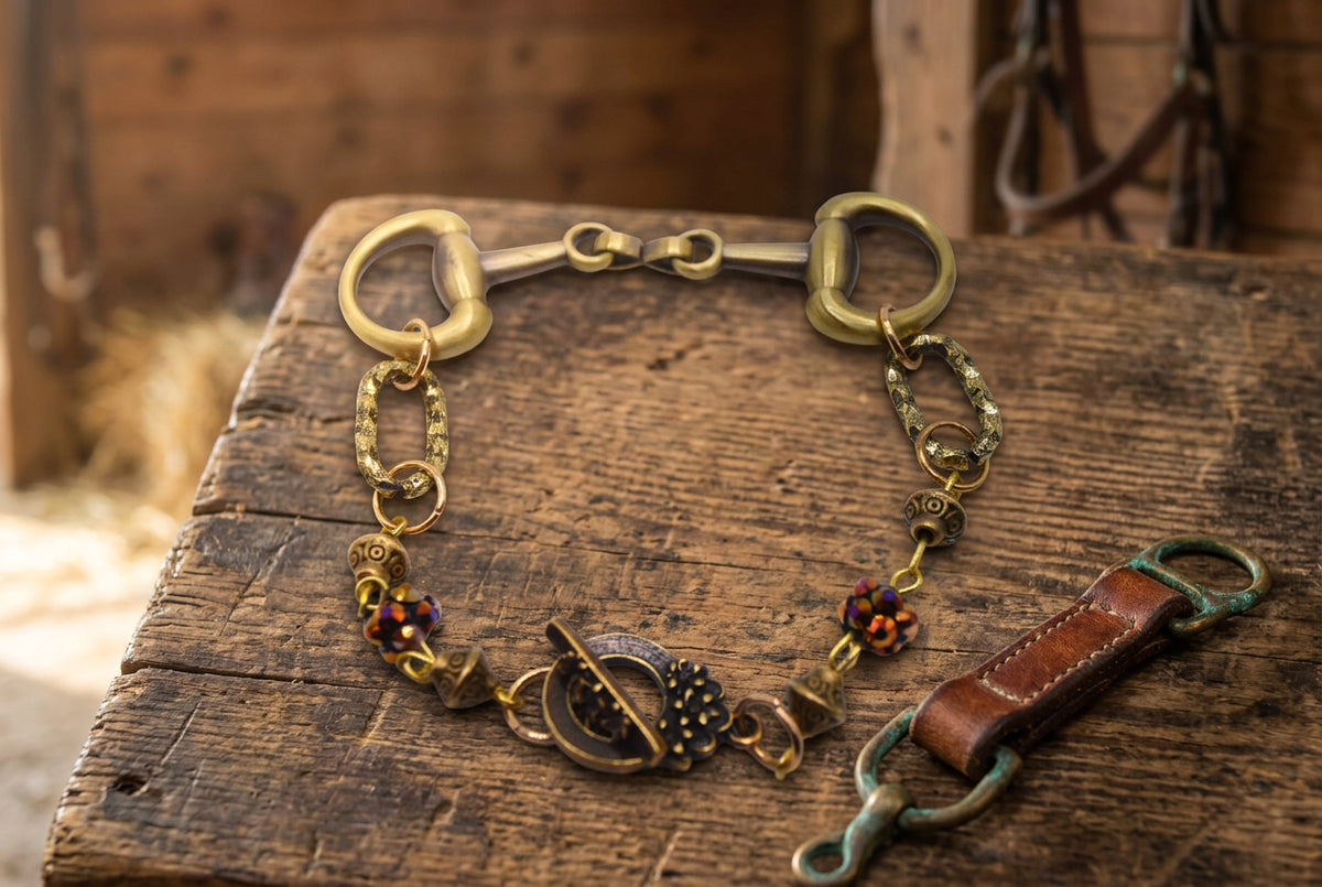An antique-style horsebit bracelet featuring a brass horsebit clasp, textured chain links, and decorative bead accents with small red and orange rhinestones, displayed on a neutral gray background.