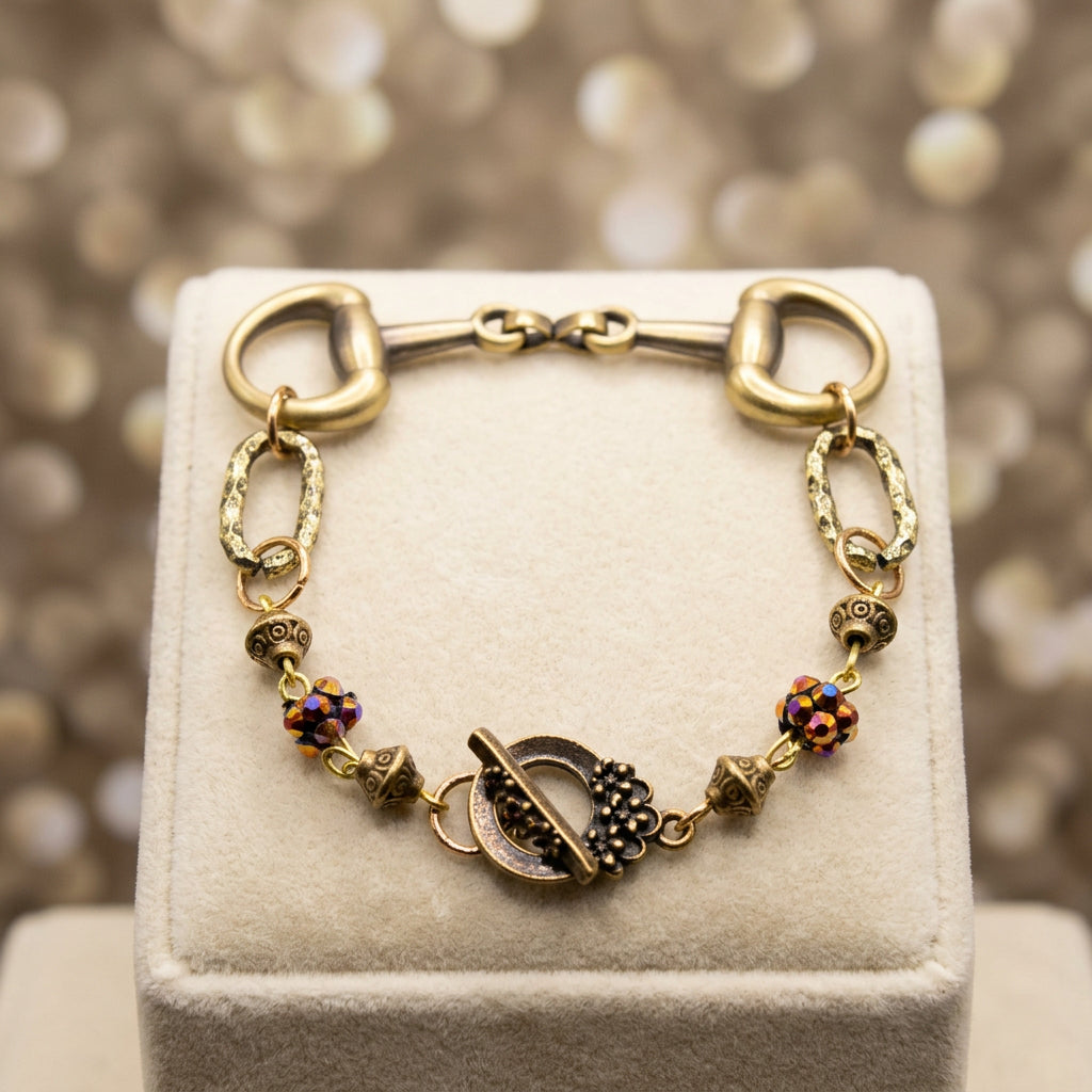 A vintage-style horse bit bracelet featuring a gold-toned horse bit clasp, chain links, decorative beads, and a toggle clasp, displayed on a neutral gray background.