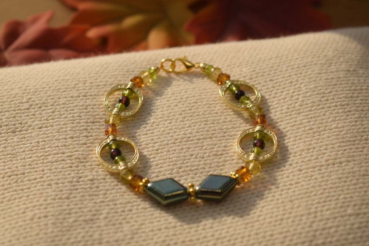 A beaded bracelet with gold-tone metal links, small faceted beads in amber and olive green, dark brown beads, and two larger iridescent blue-green diamond-shaped beads at the front, displayed on a neutral light gray background.