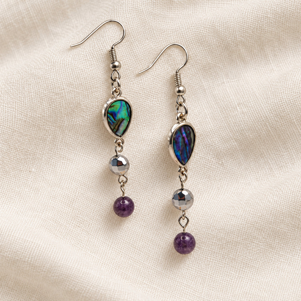 A pair of silver dangle earrings with iridescent green and blue teardrop-shaped stones, small silver beads, and dark purple round beads, hanging on a clear acrylic display stand against a light gray background.