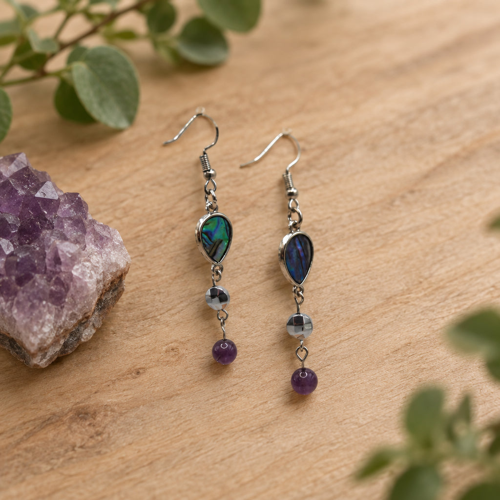 A pair of handmade dangle earrings displayed on a clear acrylic stand against a light gray background. The earrings feature teardrop-shaped iridescent abalone shell pendants with silver-toned settings, small silver beads, and dark purple round beads hanging at the bottom.