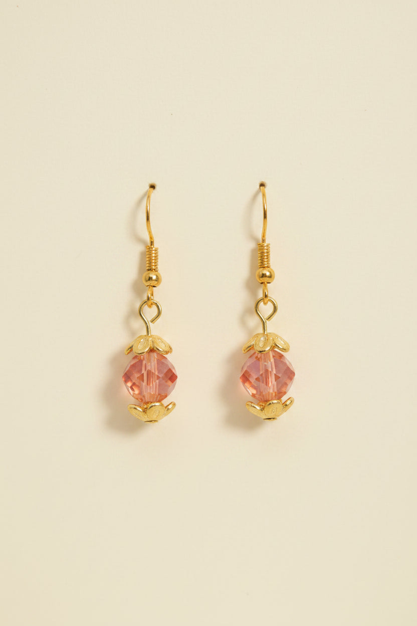 A pair of handmade dangle earrings with gold-toned metal and pinkish-orange beads, displayed on a white card labeled 'River Inspired Handcrafted Jewelry' against a plain light gray background.