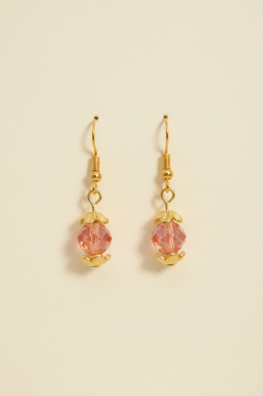 A pair of handmade dangle earrings with gold-toned metal and pinkish-orange beads, displayed on a white card labeled 'River Inspired Handcrafted Jewelry' against a plain light gray background.