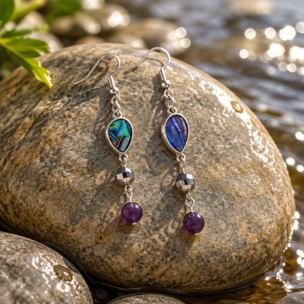 A pair of handmade dangle earrings displayed on a clear acrylic stand against a neutral gray background. The earrings feature a silver-toned fishhook ear wire, a teardrop-shaped iridescent green and blue stone, a small silver bead, a striped spacer bead, and a dangling dark purple bead.