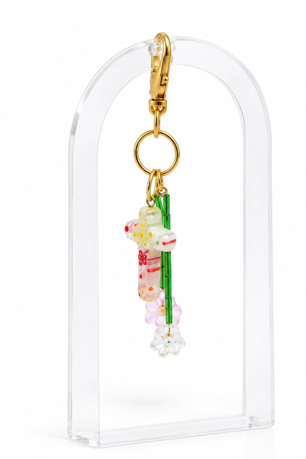 A small hanging keychain or bag charm with a gold clasp and ring, featuring a translucent pink and white cross with green and red details, displayed on a clear acrylic stand against a light gray background.